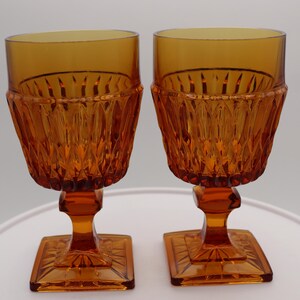May include: Two amber-colored vintage goblets. The glasses have a square base, a stem, and a textured bowl. The bowl has a diamond pattern. The goblets are made of glass and are displayed on a white surface.