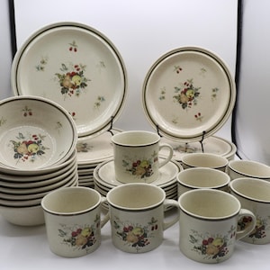 May include: A dinnerware set featuring off-white ceramic plates, bowls, and mugs decorated with a central fruit and berry motif. Each piece is accented with a dark green rim. The collection includes multiple stacked bowls, plates, and several mugs.