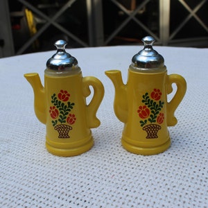 May include: Two yellow ceramic salt and pepper shakers shaped like coffee pots with silver lids and floral designs.