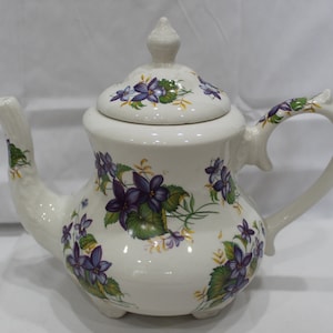 May include: White ceramic teapot with a floral design featuring purple and green violets. The teapot has a lid and a handle.