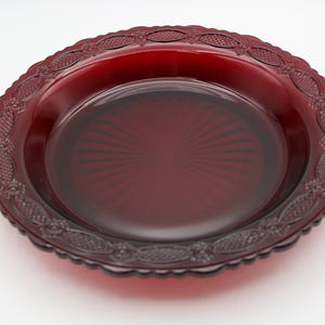May include: A ruby red glass pie plate with a decorative, scalloped edge. The plate features an embossed floral and geometric pattern around the rim. The center of the plate has a sunburst design.