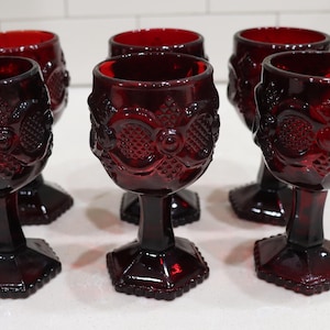 May include: A set of six deep red glass goblets featuring a raised geometric pattern on the bowl and a hexagonal base. Each drinking glass has a thick stem and a textured surface with circular and diamond-shaped embossed details in dark red glass.