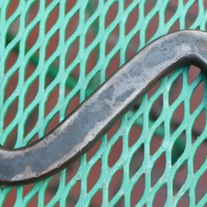 May include: A vintage, dark gray wrench with two open ends. The tool has a curved, S-shaped design and appears to be made of metal. The wrench is lying on a green, diamond-patterned surface.