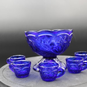 May include: A cobalt blue glass punch bowl with a pedestal base and six matching cups. The bowl and cups have a decorative, embossed pattern.