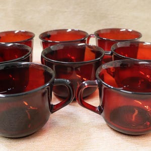 May include: Set of eight ruby red glass cups with handles. The cups have a rounded shape and a dark, transparent color. The cups are arranged in a circular pattern, showcasing their vintage design. Ideal for serving beverages.