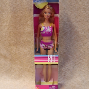 Op de afbeelding: Een Barbie-pop in een doorzichtige plastic doos. De pop heeft blond haar en draagt een tweedelige badpak met een roze, paars en wit wervelpatroon. De doos heeft het Barbie-logo en de woorden "Rio de Janeiro".