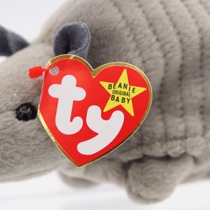 May include: A plush gray armadillo toy with a red heart-shaped tag. The tag has the words "Beanie Original Baby" and the "ty" logo. The armadillo has small gray ears and a black eye.