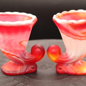 May include: Two red and white swirl glass candy dishes with a horn-shaped handle. The dishes are made of glass and have a white swirl pattern.