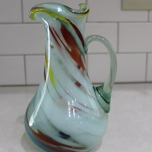 Może przedstawiać: Biały dzbanek szklany z wzorem spiralnym w odcieniach czerwieni, żółci i zieleni. Dzbanek ma szeroki otwór i uchwyt.