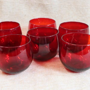 Pode incluir: Seis taças de vinho tinto sem haste. As taças são de cor vermelha escura e têm formato arredondado. São feitas de vidro e estão dispostas sobre uma superfície clara. As taças estão vazias.