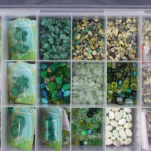 Może przedstawiać: Przezroczysty plastikowy pojemnik do przechowywania wypełniony różnymi zielonymi i białymi koralikami, kamieniami i innymi materiałami do tworzenia biżuterii. Pojemnik ma 12 przegródek i jest oznaczony etykietą "Buddies".