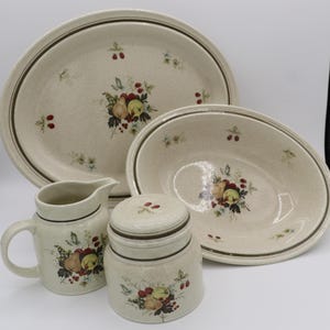 May include: A ceramic dinnerware set featuring a speckled cream base with a brown rim and a central fruit motif of pears, apples, and berries. The set includes an oval platter, an oval bowl, a lidded canister, and a small pitcher with a handle.