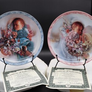 May include: Two decorative plates featuring sleeping babies surrounded by flowers and puppies. One plate has a blue border, the other a pink border. Each plate is displayed on a black metal stand, with a certificate of authenticity.