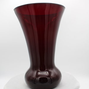 May include: A deep red glass vase with a rounded base and a flared opening. The vase is made of thick glass and has a glossy finish.
