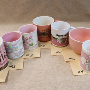 Op de afbeelding: Een assortiment keramische mokken en een grote beker in verschillende tinten roze en wit. Sommige mokken hebben bloemmotieven, vlinders en tekst zoals "Sisters and Chocolate" en "Secretary". De mokken zijn gerangschikt op een neutrale ondergrond.