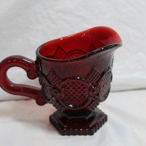 May include: A red glass creamer with a decorative pattern and a handle. The creamer has a footed base.