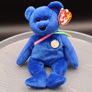 May include: A blue Beanie Baby teddy bear with a rainbow ribbon and a Ty Beanie Baby heart tag. The bear is sitting on a gray surface.