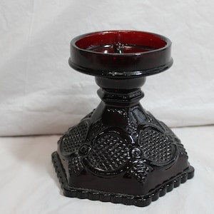 May include: A dark red glass candle holder with a square base and a round top. The base features a textured pattern with floral and geometric designs. The top has a deep red interior, and the holder is made of a glossy, reflective material.