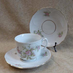 May include: A white porcelain teacup and saucer set with a matching plate. The set features a floral design with pink roses and green leaves, accented with gold trim. The teacup is on a matching saucer, and a plate is displayed behind it.