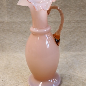 May include: A pink glass pitcher with a ruffled top and a twisted handle. The pitcher has a pearlescent finish and a clear glass base.