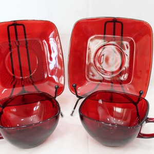 May include: Two sets of ruby red glass teacups and saucers. The cups and saucers are square shaped with a rounded bottom. The saucers have a raised center.