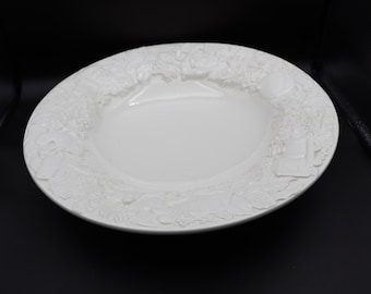 May include: A white, round serving dish with a detailed embossed border. The border features a repeating pattern of floral and architectural elements. The dish has a smooth, glossy surface and is set against a black background.