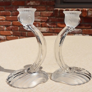 May include: A pair of clear glass candle holders featuring curved, ribbed stems and wide, scalloped bases. The top of each holder has a frosted, fluted cup designed to hold a taper candle, showcasing a blend of transparent and matte glass textures.