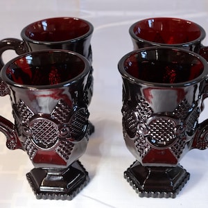 May include: Four ruby red glass mugs with ornate, textured designs. Each mug features a pedestal base, a detailed handle, and a decorative pattern on the front. The mugs are likely vintage or antique, adding a touch of elegance to any collection.