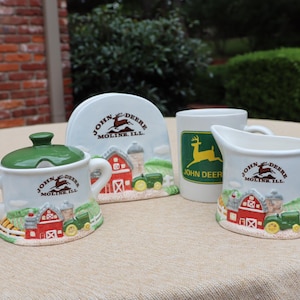 May include: A ceramic John Deere set featuring a sugar bowl with a green lid, a napkin holder, a mug with a green deer logo, and a creamer. Each piece displays a farm scene with a red barn, green tractor, and the text John Deere Moline, Ill.