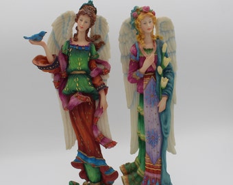 May include: Two porcelain angel figurines with wings. One angel is wearing a green, red, and gold dress with a blue bird on her hand. The other angel is wearing a purple, blue, and gold dress with a bouquet of white flowers.