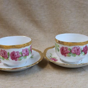 May include: Two white teacups and saucers with a gold rim and a floral rose design. The teacups have a handle and sit on matching saucers. The teacups and saucers are a vintage style.