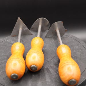 May include: Three vintage wood-handled metal tools with different shaped blades. The tools are on a black surface.