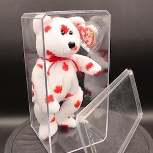 May include: A white plush bear with red maple leaf patterns, a red ribbon, and a black nose and eyes. The bear is inside a clear plastic display case with a red heart-shaped tag.