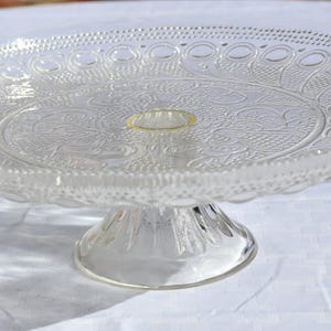 May include: Clear glass cake stand with a decorative, embossed floral pattern on the plate and a pedestal base. The rim of the plate features a series of small, circular cutouts. The stand is on a white surface.