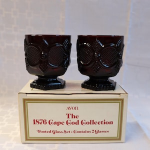 以下が含まれることがあります： Avon 1876 Cape Codコレクションのルビーレッドのフットグラス2個。グラスは装飾的なデザインで、「Footed Glass Set - Contains 2 Glasses」と書かれた箱の上に置かれています。