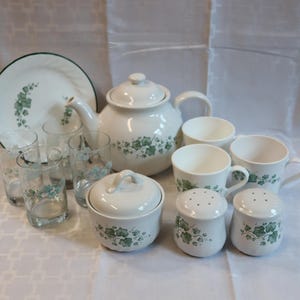 May include: A white tea set with green ivy leaf pattern. The set includes a teapot, cups, saucers, glasses, a sugar bowl, and salt and pepper shakers. The plate has a green rim.