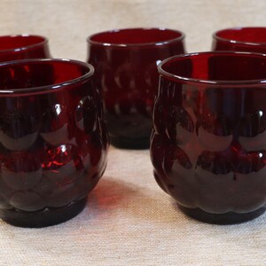 May include: Five ruby red drinking glasses. The glasses have a textured, bubble-like pattern on the exterior. The rims are smooth, and the bases are solid. The glasses are arranged on a neutral-colored surface.