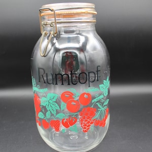 May include: A clear glass jar with a metal lid and a green and red design. The jar has the word "Rumtopf" printed on the front.