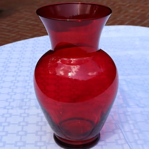May include: A tall, ruby-red glass vase with a wide, rounded body and a flared top. The vase has a glossy finish and a textured appearance. The vase is likely used for decorative purposes, such as holding flowers.