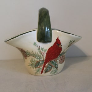 May include: A white ceramic basket with a green handle and a cardinal bird design. The basket is decorated with pine branches, pine cones, and holly berries.