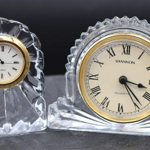 May include: Two crystal clock stands with gold-toned clock faces. The clock on the left has Roman numeral markings. The clock on the right has black markings and the word "Shannon" printed on the face.