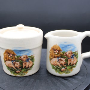 May include: Cream-coloured ceramic sugar bowl with lid and creamer set. Each piece features a pastoral scene with pigs wearing blue bows, a barn, and a fence. The set is designed for serving sugar and cream.