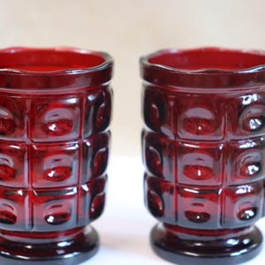 May include: Two ruby red vintage drinking glasses. Each glass has a textured, square pattern around the body and a scalloped rim. The glasses are made of a dark red glass and sit on a small base.