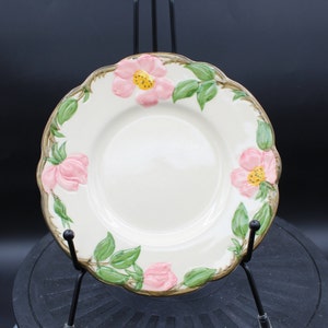 May include: A white ceramic plate with a scalloped edge and a floral design. The plate features pink roses with yellow centers and green leaves.