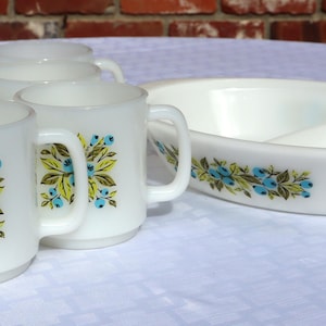 Op de afbeelding: Een witte vintage servies set met een bloemen design. De set bevat zes mokken en een verdeelde serveerschaal. Het bloemenpatroon heeft blauwe, gele en groene accenten. De mokken hebben een handvat en een afgeronde basis.