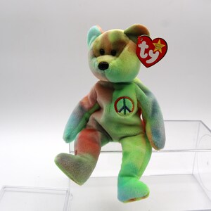 May include: A colorful plush bear with a peace sign on its chest and a TY tag. The bear features a tie-dye pattern in shades of green, blue, orange, and brown. The bear is sitting in a clear plastic container.