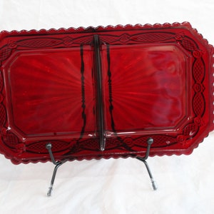Op de afbeelding: Een rechthoekige, robijnrode glazen schaal met een geschulpte rand en decoratieve details. De schaal wordt ondersteund door een zwarte metalen standaard. Het glas heeft een zonnestraalpatroon dat vanuit het midden uitstraalt.