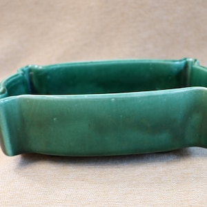 Op de afbeelding: Een rechthoekige, smaragdgroene keramische plantenbak met een geschulpte rand. De plantenbak heeft een glanzende afwerking en een licht gebogen vorm. Het algehele ontwerp is eenvoudig en elegant, geschikt voor het tentoonstellen van kleine planten of bloemen.