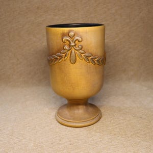 May include: A decorative, antique-style goblet with a pedestal base. The goblet is a light tan color with a darker tan decorative floral and leaf design around the top. The interior is black.
