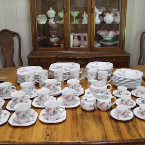 May include: A complete vintage floral china set, including cups, saucers, plates, and serving pieces. The set features a white background with pink and green floral designs. The arrangement is on a wooden table, with a wooden cabinet and chairs in the background.